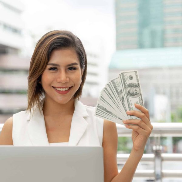 Beautiful girl smiling in business woman clothes  using laptop computer and show money US dollar bills in hand , urban city background